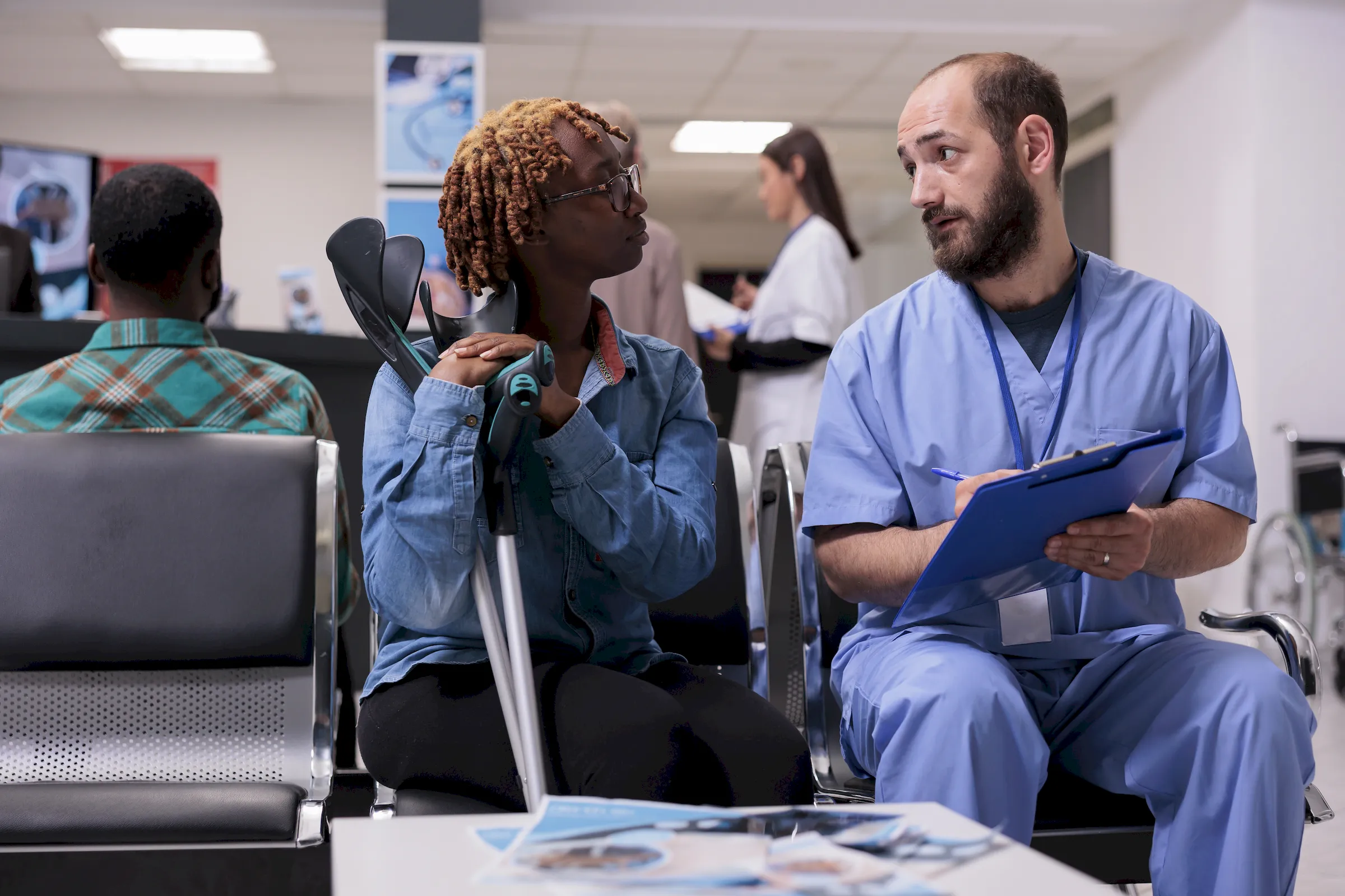 A clinician with a clipboard talking to a patient with crutches in a waiting area.
